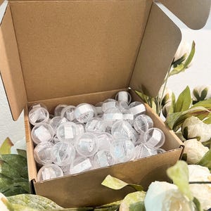 May include: A cardboard box filled with clear plastic spheres, some with the word "Party" printed on them. The box is surrounded by white flowers and green leaves, creating a decorative display.