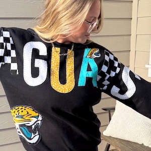 May include: A black long-sleeve sweatshirt featuring the word JAGUARS spelled out in large, multi-patterned fabric letters across the chest and sleeves. The design includes a large jaguar head patch on the lower front and checkered fabric accents.
