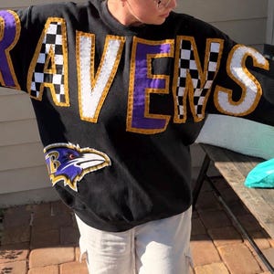 May include: A black long-sleeve sweatshirt featuring the word RAVENS in large, stitched-on letters across the chest. The letters alternate between solid purple and black-and-white checkered patterns, outlined in gold. A Baltimore Ravens logo is below.