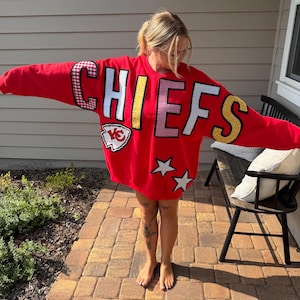 May include: A bright red crewneck sweatshirt featuring the word CHIEFS in large, multi-patterned, and sequined block letters across the chest. Below the text is a white and red arrowhead logo patch and two white star-shaped patches on the lower front.