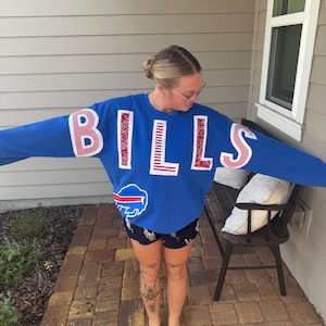 May include: A royal blue long-sleeve sweatshirt featuring the word BILLS in large, white-bordered letters with red and white patterned fabric fill. Below the text is a red, white, and blue Buffalo Bills team logo patch on the front.