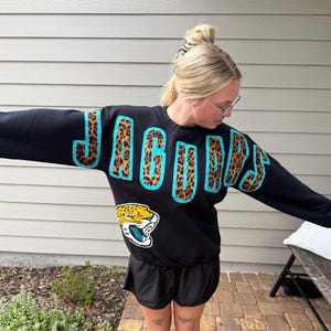 May include: A black long-sleeve sweatshirt featuring the word JAGUARS in large, leopard-print letters outlined in bright teal. Below the text, a graphic of a snarling jaguar head with yellow, black, and white fur and a teal mouth is displayed.