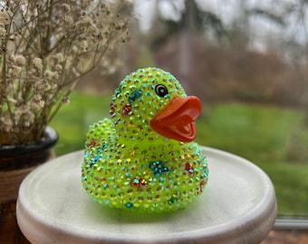 May include: A lime green rubber ducky covered in iridescent rhinestones. The duck has an orange beak and a black eye.