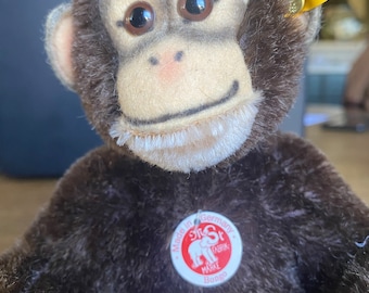 Puede incluir: Un mono de peluche marrón con una cara blanca y grandes ojos marrones. El mono lleva una etiqueta roja que dice "Steiff, Fabrik, Marke, Bongo" y una etiqueta amarilla que dice "Made in Germany, Steiff, Knopf im Ohr".