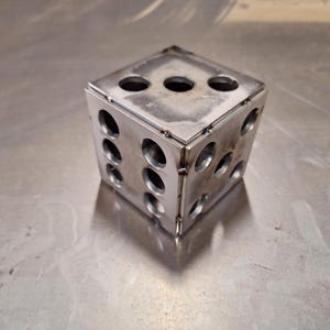 May include: A handcrafted, metallic dice with rounded holes representing the numbers. The cube is made of welded metal and sits on a reflective surface. The dice is a unique decorative item.