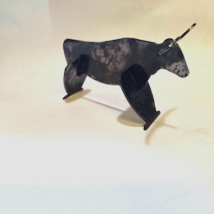 Op de afbeelding: Een zwarte metalen sculptuur van een stier met een getextureerd lichaam en vier poten. De stier heeft een hoorn en staat op een wit oppervlak. De sculptuur is een uniek kunstwerk.