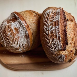 Könnte beinhalten: Zwei frisch gebackene Brote mit einem Blattdesign auf der Oberseite. Das Brot liegt auf einem Holzbrett.