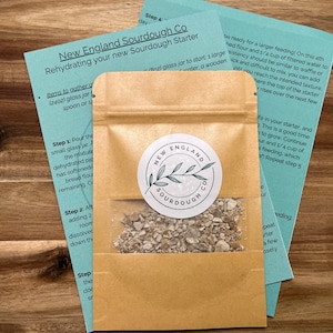 May include: A brown paper packet with a white label that says "New England Sourdough Co." and contains a dehydrated sourdough starter. The packet is sitting on a wooden surface with instructions for rehydrating the starter.