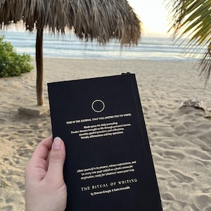 May include: A black journal with gold lettering that reads "This is the journal that will inspire you to write." The journal has a circle symbol on the front cover and the title "The Ritual of Writing" by Shawna Krueger & Katie McAuliffe.