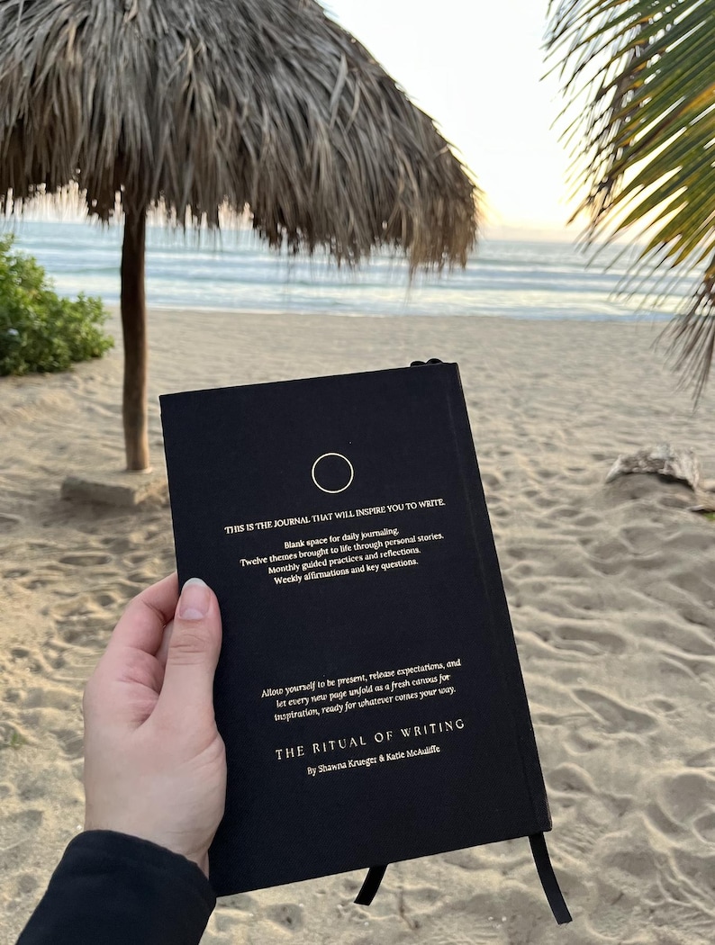 May include: A black journal with gold lettering that reads "This is the journal that will inspire you to write." The journal has a circle symbol on the front cover and the title "The Ritual of Writing" by Shawna Krueger & Katie McAuliffe.