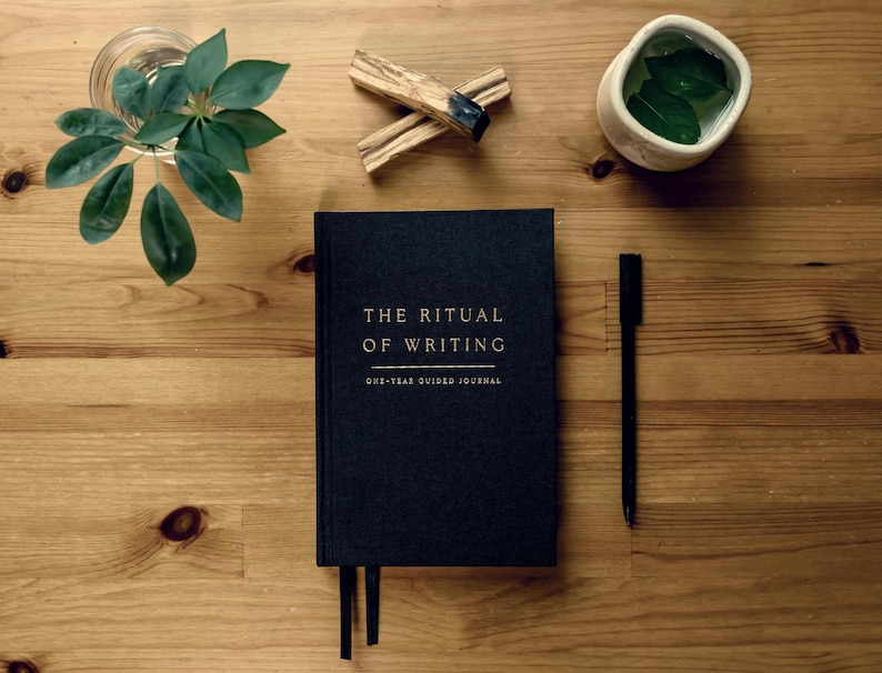 The Ritual of Writing journal on top of a desk with a pen, plant, palo santo, and cup of tea
