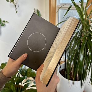 May include: A black hardcover book with gold-gilded edges is open to a page with a circle design. The book is being held by a person's hands.