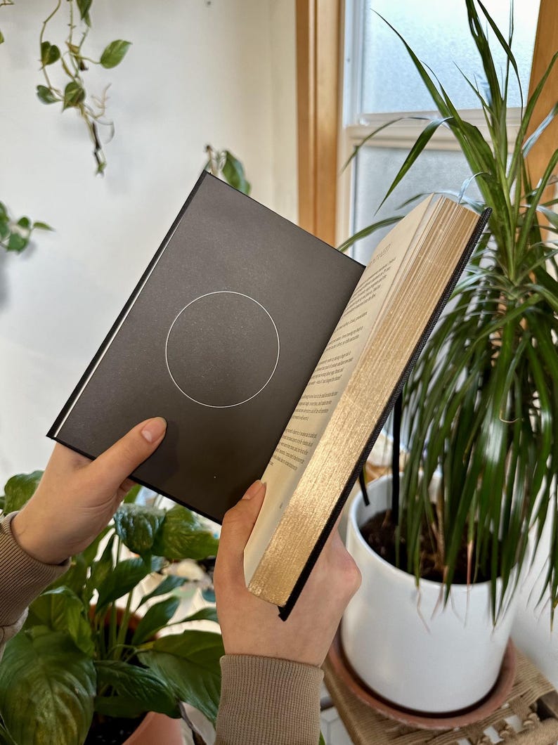 May include: A black hardcover book with gold-gilded edges is open to a page with a circle design. The book is being held by a person's hands.