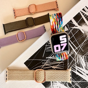 May include: Five different Apple Watch bands, including a rainbow woven band, a beige woven band, a lavender woven band, a dark green woven band, and a light pink woven band. The rainbow band is on a silver Apple Watch with a black face.