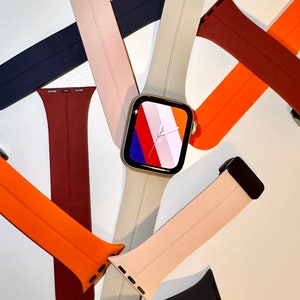 Op de afbeelding: Een assortiment van kleurrijke siliconen horlogebandjes voor een Apple Watch. De bandjes zijn in tinten oranje, rood, beige en zwart. De wijzerplaat van het horloge is zichtbaar met een kleurrijk abstract ontwerp.