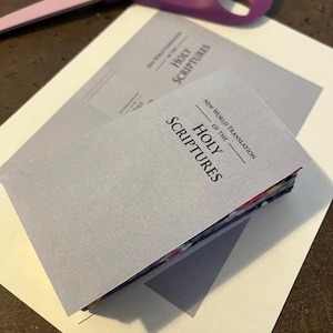 以下が含まれることがあります： 「NEW WORLD TRANSLATION OF THE HOLY SCRIPTURES」の文字が入った、2冊の薄い灰色の小冊子が白い紙の上に重ねられています。紫色のハサミが上に置かれています。小冊子の端にはカラフルなページが見えます。