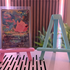 以下が含まれることがあります： ピンクと水色のミニチュアイーゼルが2つ、ポケモンのトレーディングカードの隣にあります。カードにはピンク色の漫画のキャラクターと日本語のテキストが描かれています。イーゼルはプラスチック製で、小さなアイテムを保持するように設計されています。
