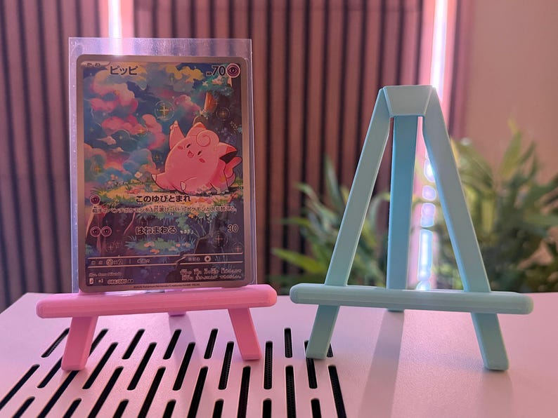 以下が含まれることがあります： ピンクと水色のミニチュアイーゼルが2つ、ポケモンのトレーディングカードの隣にあります。カードにはピンク色の漫画のキャラクターと日本語のテキストが描かれています。イーゼルはプラスチック製で、小さなアイテムを保持するように設計されています。