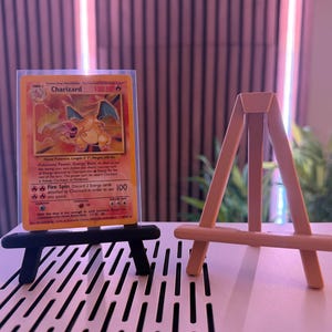 May include: A Charizard Pokémon trading card displayed on a black miniature easel. The card features a fiery orange and yellow dragon illustration. A second, natural wood-colored easel is also present.