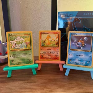 以下が含まれることがあります： ミニチュアイーゼルに飾られた、フシギダネ、ヒトカゲ、ゼニガメの3枚のポケモンカード。カードには、キャラクターのカラフルなイラストが描かれています。ゲンガーのフィギュアとポケモンのエリートトレーナーボックスが背景にあります。