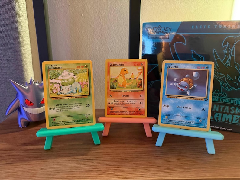 以下が含まれることがあります： ミニチュアイーゼルに飾られた、フシギダネ、ヒトカゲ、ゼニガメの3枚のポケモンカード。カードには、キャラクターのカラフルなイラストが描かれています。ゲンガーのフィギュアとポケモンのエリートトレーナーボックスが背景にあります。