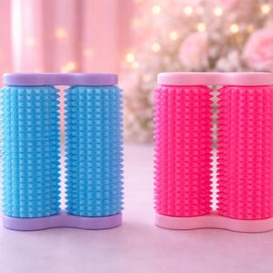 Op de afbeelding: Twee haarrollers, één blauw en één roze, elk met een textuuroppervlak. De rollers zijn aan de basis en bovenkant verbonden. De achtergrond is zachtroze met vage bloemen en lichtjes.