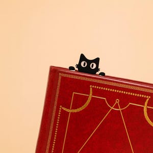 以下が含まれることがあります： 赤い本の上から黒猫のブックマークが覗いています。本には金色の装飾と幾何学模様があります。背景は単色の明るいピーチ色です。