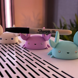 May include: Three small, colourful, 3D printed figurines shaped like crabs. The crabs are white, purple, and light blue. A black Sharpie fine point permanent marker is lying on top of the crabs.
