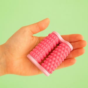 Puede incluir: Un rodillo para pies rosa con superficies texturizadas, sostenido en una mano sobre un fondo verde. El rodillo tiene dos secciones con protuberancias y crestas redondeadas y elevadas. El marco es de un tono rosa más claro.