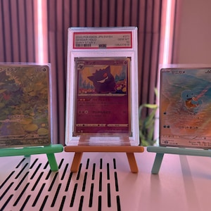 以下が含まれることがあります： ミニチュアイーゼルに飾られた3枚のポケモンカード。カードには様々なキャラクターと日本語のテキストが描かれています。中央のカードは、「2020 Pokemon JPN SWSH Gengar Hold Shiny Star V」と書かれた保護ケースに入っています。