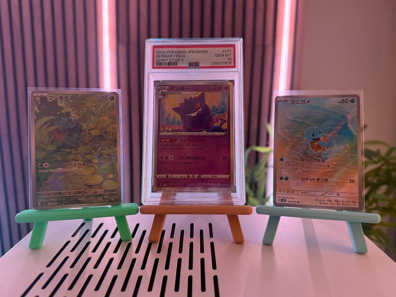 以下が含まれることがあります： ミニチュアイーゼルに飾られた3枚のポケモンカード。カードには様々なキャラクターと日本語のテキストが描かれています。中央のカードは、「2020 Pokemon JPN SWSH Gengar Hold Shiny Star V」と書かれた保護ケースに入っています。