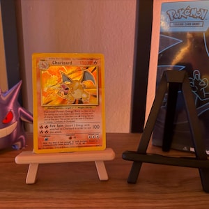 以下が含まれることがあります： 小さな木製イーゼルに飾られた、Charizardのポケモンカード。燃えるようなオレンジ色のドラゴンのイラストが描かれています。紫色のゲンガーのフィギュアとポケモンカードバインダーが背景にあります。