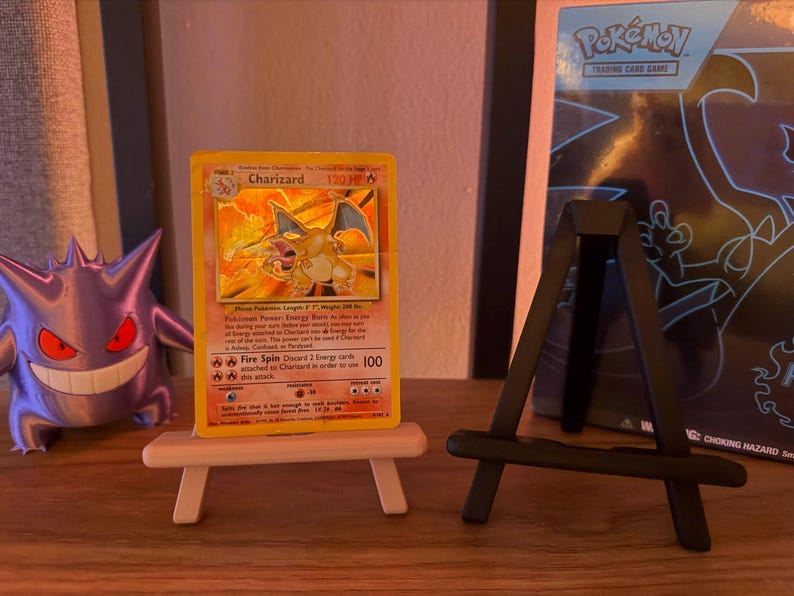 以下が含まれることがあります： 小さな木製イーゼルに飾られた、Charizardのポケモンカード。燃えるようなオレンジ色のドラゴンのイラストが描かれています。紫色のゲンガーのフィギュアとポケモンカードバインダーが背景にあります。