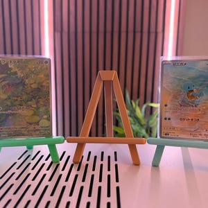 以下が含まれることがあります： 小さなイーゼルに飾られた2枚のポケモンカード。カードは保護スリーブに入っています。イーゼルはオレンジ色、他の2つは緑色と青色です。カードには日本語のテキストとカラフルなイラストが描かれています。
