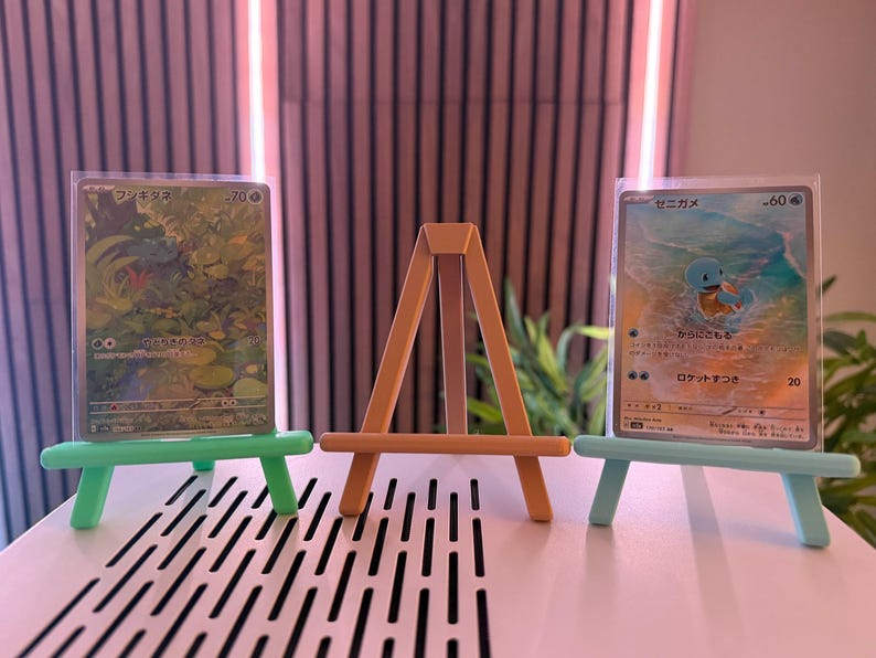 以下が含まれることがあります： 小さなイーゼルに飾られた2枚のポケモンカード。カードは保護スリーブに入っています。イーゼルはオレンジ色、他の2つは緑色と青色です。カードには日本語のテキストとカラフルなイラストが描かれています。