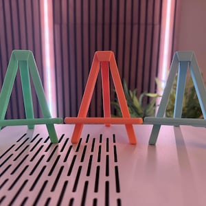 以下が含まれることがあります： ミントグリーン、コーラル、ライトブルーの3色の小さなイーゼルが並んでいます。各イーゼルは、水平なサポートバーを備えた三角形のAフレームデザインです。イーゼルは無地のマットな素材でできており、ぼやけた背景に配置されています。