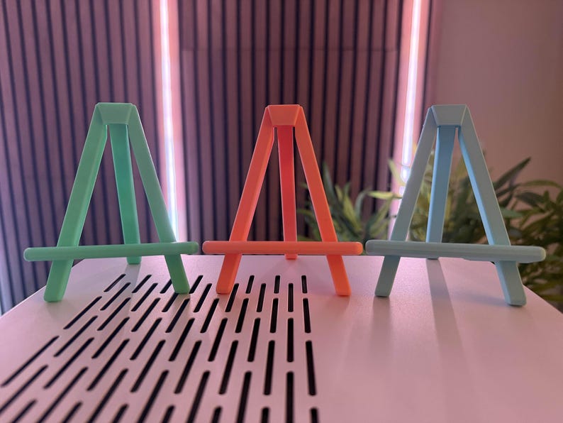 以下が含まれることがあります： ミントグリーン、コーラル、ライトブルーの3色の小さなイーゼルが並んでいます。各イーゼルは、水平なサポートバーを備えた三角形のAフレームデザインです。イーゼルは無地のマットな素材でできており、ぼやけた背景に配置されています。