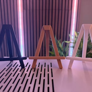 以下が含まれることがあります： 黒、ベージュ、白の3つの小さな木製イーゼルが表示されています。 イーゼルは並んで配置され、シンプルなAフレームデザインです。 パターンのある白い表面に置かれ、背景はぼやけています。
