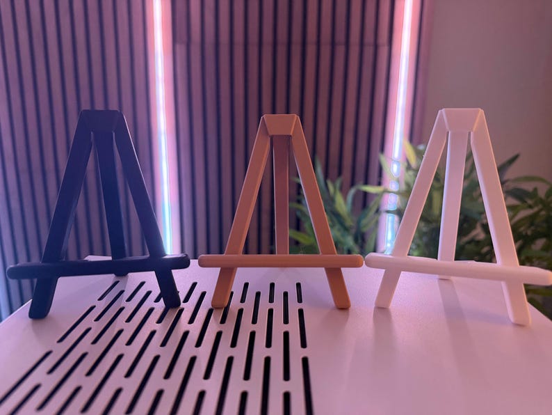 以下が含まれることがあります： 黒、ベージュ、白の3つの小さな木製イーゼルが表示されています。 イーゼルは並んで配置され、シンプルなAフレームデザインです。 パターンのある白い表面に置かれ、背景はぼやけています。