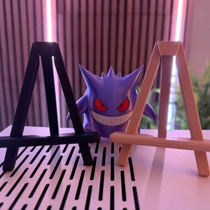 以下が含まれることがあります： 黒とタンの2つの小さな木製イーゼルが並んでおり、背景には紫色のゲンガーのフィギュアがあります。ゲンガーは赤い目と大きな笑顔をしています。イーゼルは白い表面にあります。