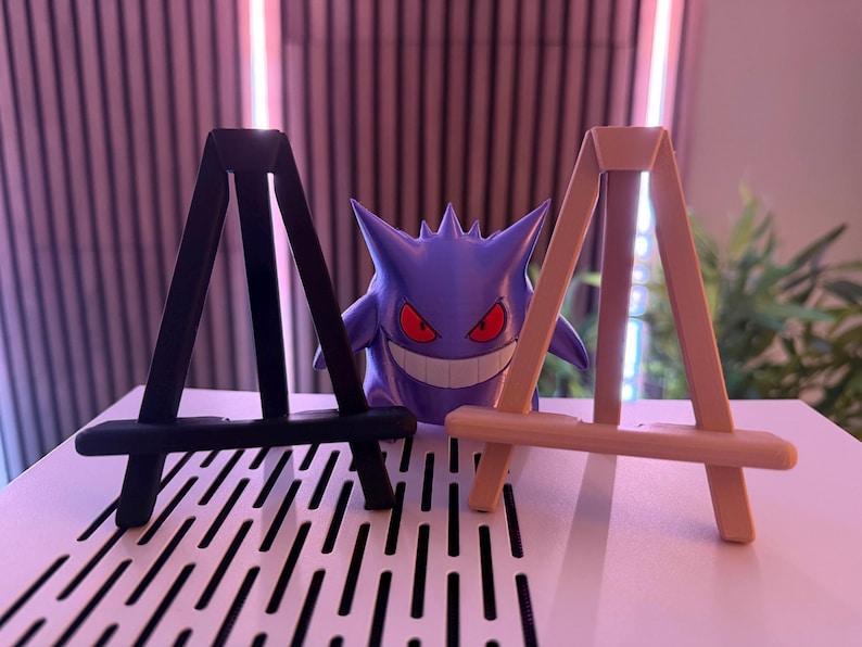 以下が含まれることがあります： 黒とタンの2つの小さな木製イーゼルが並んでおり、背景には紫色のゲンガーのフィギュアがあります。ゲンガーは赤い目と大きな笑顔をしています。イーゼルは白い表面にあります。