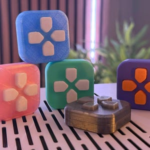 Puede incluir: Cinco botones cuadrados de diferentes colores con un pad direccional. Los botones son azul claro, rosa, verde, gris y morado. Los botones tienen pads direccionales blancos, grises y naranjas.