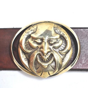 May include: A gold-toned belt buckle with a detailed design of a grinning devil's face. The buckle is oval-shaped and has a smooth, polished finish.