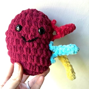 Puede incluir: Un riñón de felpa hecho a mano, de color burdeos, con ojos de seguridad negros y una sonrisa cosida. El riñón tiene detalles de ganchillo rojos, azules y amarillos. La felpa está sujeta por una mano.