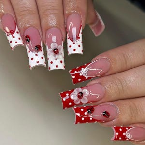 May include: A set of long, square-shaped artificial nails. The nails feature a white base with red polka dots, red tips, and a clear pink base. Some nails have 3D white flowers with red centers and ladybug accents. Other nails have white bow designs.