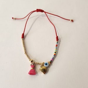 May include: A red string bracelet with a gold heart charm, a blue evil eye charm, and a pink tassel. The bracelet is made with small gold beads and colorful beads.
