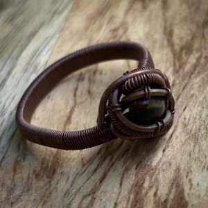 May include: A copper wire ring with a brown gemstone set in the center. The ring is made of coiled wire and has a rustic, handmade look.