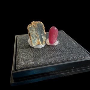 May include: Two gemstones on a black display stand. The first gemstone is a dark gray, green, and brown crystal. The second gemstone is a bright pink oval.