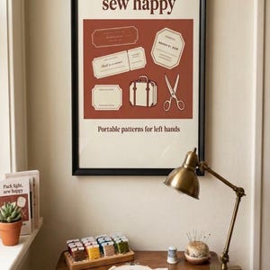 May include: A framed wall poster with a dark brown border featuring the text "Pack light, sew happy" and "Portable patterns for left hands" above and below a rust-colored graphic containing illustrations of vintage tickets, a suitcase, and scissors.