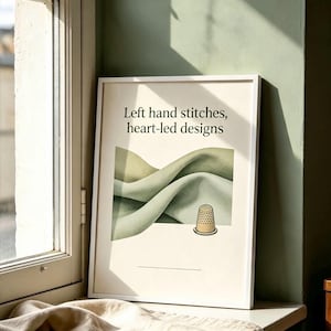 May include: A white framed wall art print featuring the text Left hand stitches, heart-led designs in black serif font. Below the text is an illustration of soft, sage green fabric folds and a single metallic gold sewing thimble.